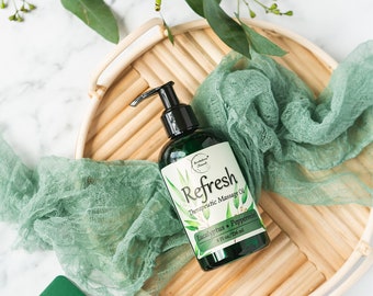 May include: A green glass bottle of Refresh Therapeutic Massage Oil with a black pump top. The label is white with green text and a green leaf design. The bottle is sitting on a woven bamboo tray with a green cloth.