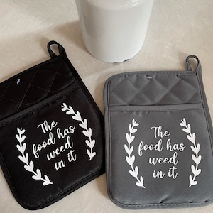 May include: Two quilted pot holders, one black and one gray, with the text "The food has weed in it" in white script. Each pot holder has a pocket and a hanging loop. A white pitcher with flowers is in the background.