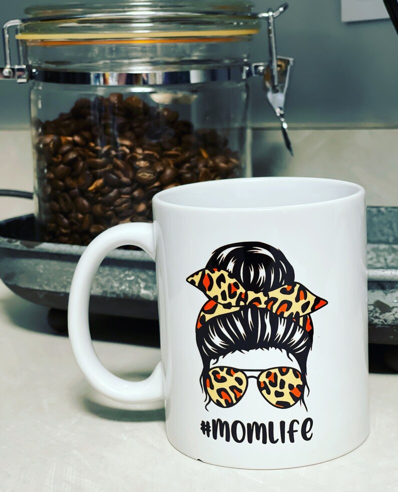 May include: White ceramic coffee mug featuring a graphic of a woman's messy bun hairstyle with leopard print sunglasses and a matching bow. The mug has the text "#momlife" in black.