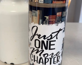 May include: A stainless steel tumbler with a wraparound design of bookshelves filled with books. The tumbler has the text "Just One More Chapter" in black script. The lid is blue.