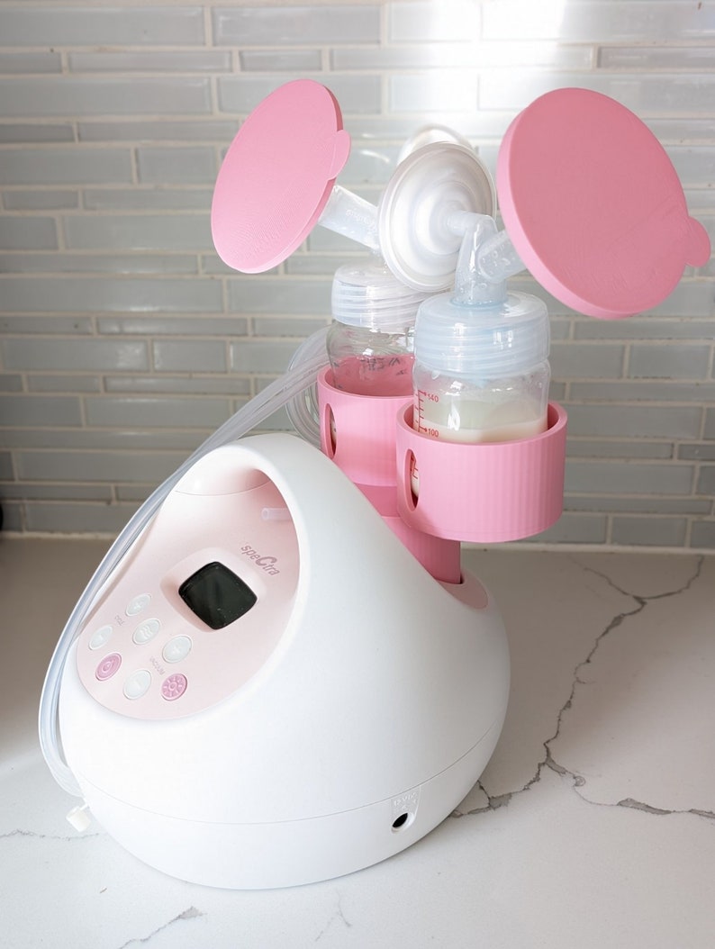 May include: A white electric breast pump with a digital display and pink accents. The pump has two bottles attached, each with a pink lid and a clear silicone funnel.