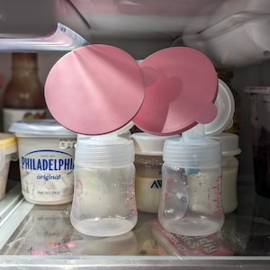 May include: Two pink silicone breast pump flanges are placed on top of two clear glass baby bottles with white lids. The bottles are filled with a white liquid and have measurement markings on the side. The bottles are sitting on a shelf in a refrigerator. The text "Humidity Control" is visible on the bottom of the image.
