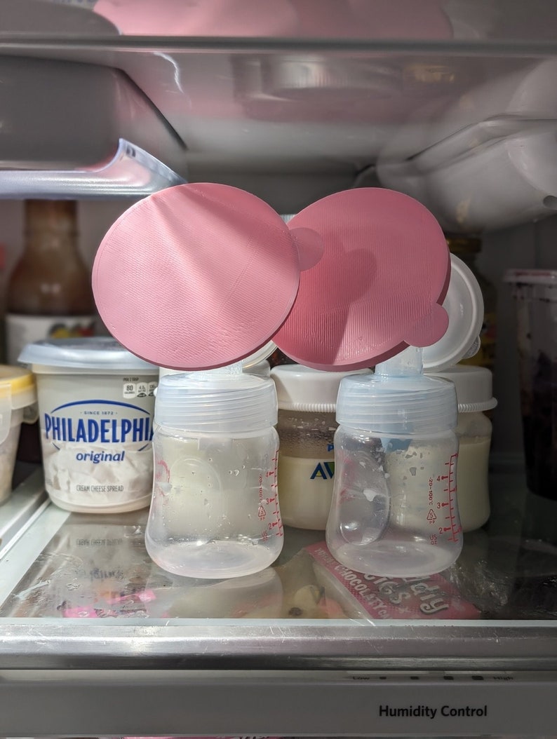 May include: Two pink silicone breast pump flanges are placed on top of two clear glass baby bottles with white lids. The bottles are filled with a white liquid and have measurement markings on the side. The bottles are sitting on a shelf in a refrigerator. The text "Humidity Control" is visible on the bottom of the image.