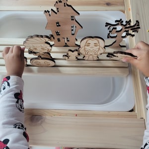 Könnte beinhalten: Ein Kind spielt mit einem Satz hölzerner Halloween-Spielzeug. Das Spielzeug umfasst ein Haus, einen Baum, einen Kürbis und zwei Figuren. Das Spielzeug befindet sich auf einer Holzoberfläche.