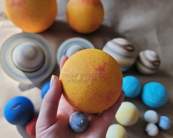 May include: A selection of handcrafted planet models, including the sun, moon, and other planets. The sun is a large, textured yellow sphere with red accents. The text "The Sun and Moon in Medium set" is visible. Other planets are blue, brown, and white.