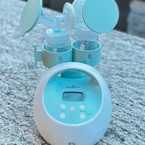 May include: A white and light blue electric breast pump with a digital display and buttons labeled "cycle" and "vacuum". The pump has two bottles attached with blue caps.