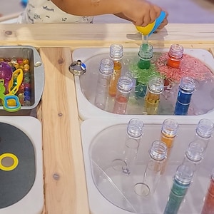 May include: A wooden activity table with multiple compartments for sensory play. One compartment has colorful beads, another has clear tubes filled with colored water, and a third has a black tray with colorful rings.