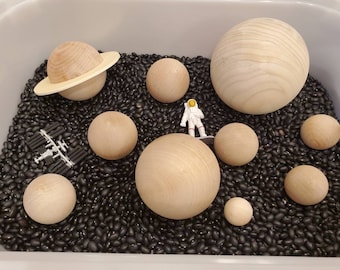May include: A space-themed sensory bin with black beans, wooden planets, a toy astronaut, and a toy space shuttle.