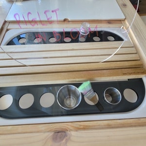 May include: A wooden art station with a clear plastic top and a black and white paint tray. The tray has circular openings for cups and a brush. The words "PIGLET IN BLOOM" are written on the top.