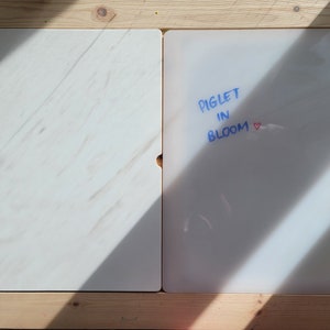 Op de afbeelding: Twee rechthoekige witte platen in een lichtgekleurd houten frame. De linkerplaat heeft een subtiel marmerachtig patroon. De rechterplaat bevat de blauwe tekst PIGLET IN BLOOM, gevolgd door een klein rood hartje op het oppervlak.