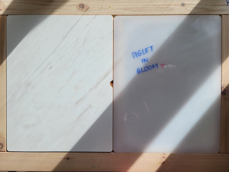 Op de afbeelding: Twee rechthoekige witte platen in een lichtgekleurd houten frame. De linkerplaat heeft een subtiel marmerachtig patroon. De rechterplaat bevat de blauwe tekst PIGLET IN BLOOM, gevolgd door een klein rood hartje op het oppervlak.
