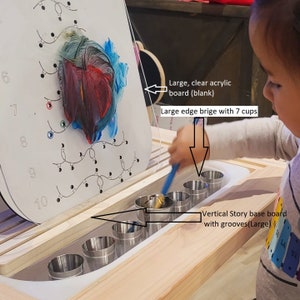 May include: A child's art station with a large, clear acrylic board featuring colorful paint strokes. The station includes a wooden base with a series of metal cups and a vertical story base board with grooves.