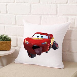 Puede incluir: Una almohada blanca con un coche de carreras de dibujos animados rojo impreso en ella. El coche tiene una lengua grande que sobresale y est&aacute; mirando al espectador.