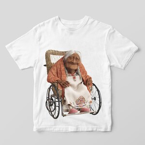 Puede incluir: Camiseta blanca con una imagen colorida de una mujer mayor sentada en una silla de ruedas de mimbre. La mujer lleva un vestido blanco con bordados florales y una chal roja.
