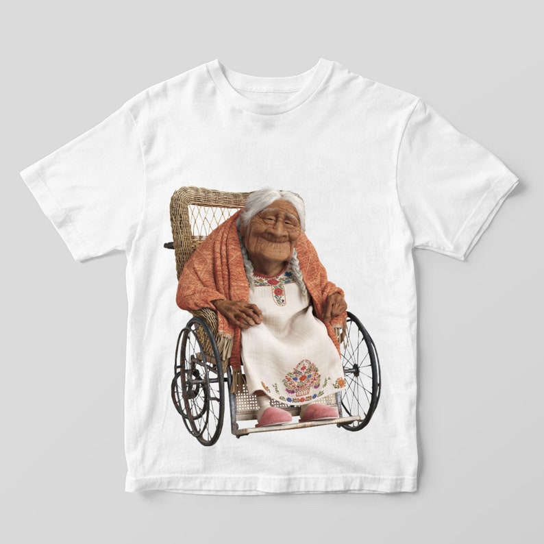 Puede incluir: Camiseta blanca con una imagen colorida de una mujer mayor sentada en una silla de ruedas de mimbre. La mujer lleva un vestido blanco con bordados florales y una chal roja.
