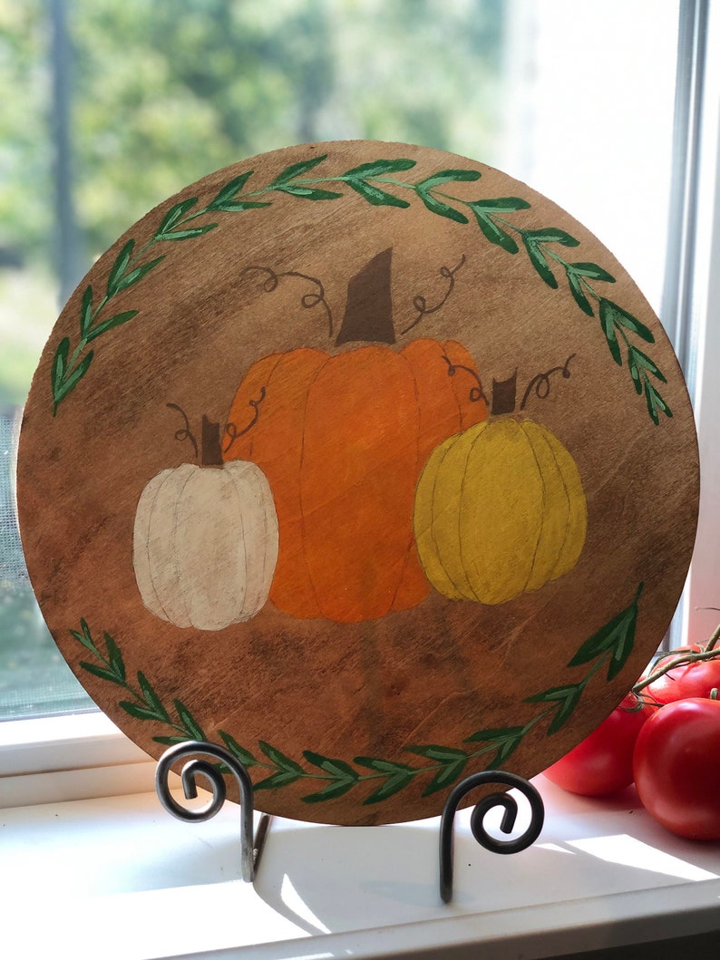 May include: A round wooden sign with a painted wreath of green leaves around the edge. Three pumpkins are painted in the center, one orange, one white, and one yellow.
