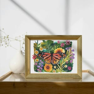 May include: A rectangular gold-toned frame containing a colorful painting of a monarch butterfly centered among various green leaves, yellow sunflowers, and small pink and purple flowers. The artwork features a vibrant botanical theme.