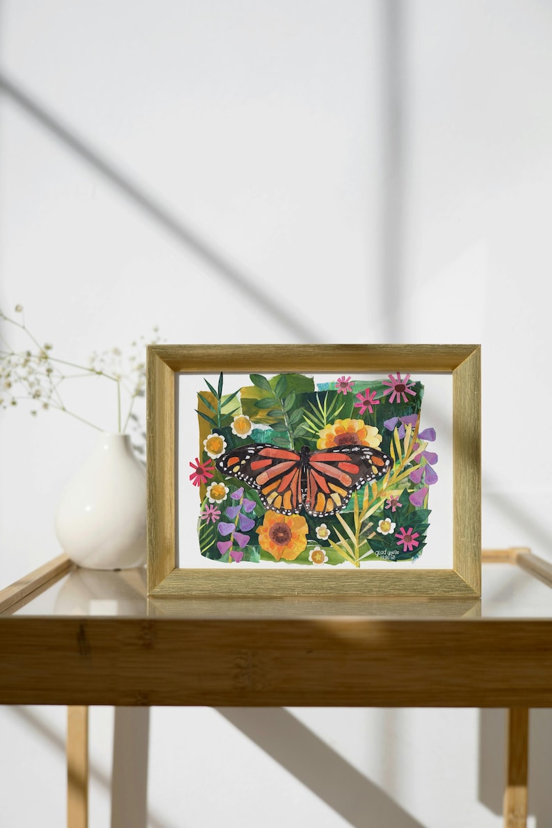 May include: A rectangular gold-toned frame containing a colorful painting of a monarch butterfly centered among various green leaves, yellow sunflowers, and small pink and purple flowers. The artwork features a vibrant botanical theme.