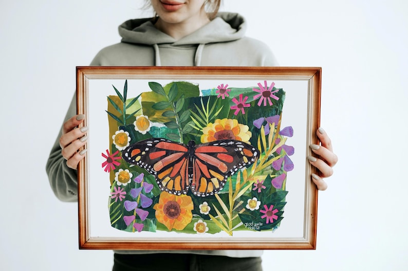 May include: A rectangular wooden frame containing a collage-style artwork of a monarch butterfly centered among various green leaves and colorful flowers in shades of yellow, pink, and purple. The text "glad you're here" is visible in the bottom right.