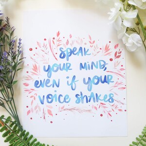 Può includere: Una stampa rettangolare bianca con il testo blu scritto a mano "Speak your mind, even if your voice shakes" circondato da una corona decorativa di foglie, steli e piccoli punti dipinti in rosa e rosso su uno sfondo bianco.