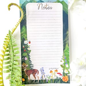 May include: A rectangular notepad with horizontal lines and the word Notes printed at the top. The border features a forest scene with green mountains, pine trees, a brown deer, red strawberries, white and orange flowers, and red mushrooms.