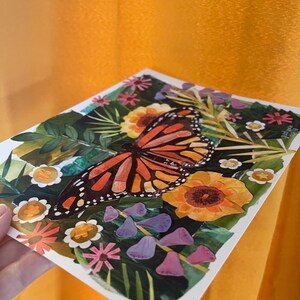 May include: A square art print featuring a central monarch butterfly with orange and black wings surrounded by a collage of green leaves, yellow sunflowers, small white flowers with gold centers, and purple bell-shaped blossoms on a dark background.
