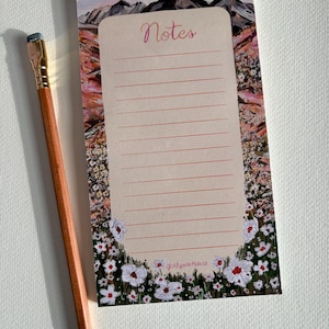 May include: A rectangular notepad with a light beige center featuring the word Notes in cursive script and horizontal lines. The border displays a painted landscape of mountains and a field of white flowers with red centers. A wooden pencil lies beside it.