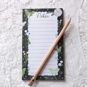 May include: A notepad with a black background, adorned with illustrations of butterflies, flowers, and foliage. The word "Notes" is written at the top. A wooden pencil rests diagonally across the notepad, ready for use.