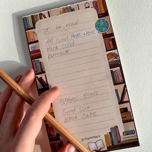 May include: A rectangular notepad with a bookshelf illustration featuring colorful books, a globe, and a clock. The cream-colored lined paper has handwritten text including To be read, All Good People Here, Buck Wild, Burnout, October Reads, Good Dirt, Black Cake, and the brand name gladyourehere.co.