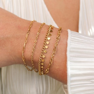 May include: A collection of gold bracelets in various styles adorn a wrist. The bracelets include a paperclip chain, a delicate chain, a chain with small circular charms, and a curb chain. The bracelets are layered and worn against a white, pleated fabric.