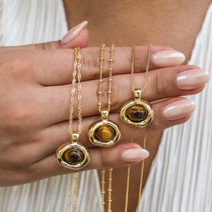 Op de afbeelding: Gouden kettingen met ovale tijgeroog hangers. De hangers zijn gezet in een getextureerde rand en hangen aan fijne kettingen. De sieraden worden gepresenteerd tegen een witte achtergrond, wat de warme tinten benadrukt.