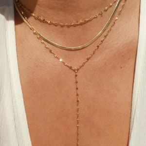 May include: Three gold necklaces layered together. The top necklace is a delicate chain, the middle is a snake chain, and the bottom is a Y-shaped necklace with a long, dangling chain. The necklaces are worn against a light background.