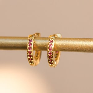May include: A pair of gold-colored hoop earrings, each encrusted with small, vibrant red gemstones. The earrings are displayed on a gold-colored bar against a soft, blurred background. The gemstones are evenly spaced around the hoops.