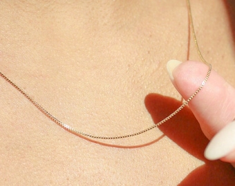 Könnte beinhalten: Eine zarte goldene Halskette mit feinem, quadratischem Kettendesign. Die Halskette wird von einer Hand mit langen, polierten Nägeln gehalten. Die Kette liegt auf der Haut und unterstreicht ihren subtilen Glanz und minimalistischen Stil.