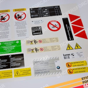 May include: A sheet of car stickers with various warning labels and safety information. Includes labels with the text "ACHTUNG WARNING", "AIRBAG", and "do not tow or lash". Also features red triangles and a barcode.