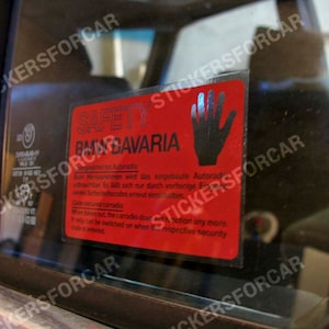 May include: A red and black safety sticker with the text "SAFETY" and "BMW BAVARIA" in white. The sticker also includes a hand symbol with a red background and a black hand.