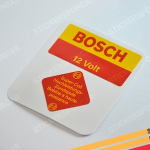 Puede incluir: Dos pegatinas rojas y amarillas para un coche. La pegatina más grande dice "BOSCH 12 Volt" y la pegatina más pequeña dice "Super-Coil Hochleistungs- Zündspule Bobine a haute puissance".