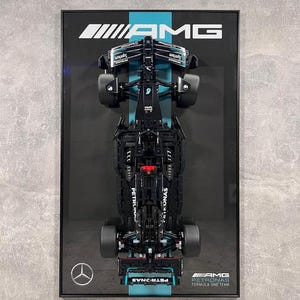 Puede incluir: Un modelo de coche de carreras de Fórmula 1 negro y azul enmarcado en un marco negro. El coche está boca abajo con las ruedas hacia arriba. El marco tiene las palabras "AMG PETRONAS FORMULA ONE TEAM" en la parte inferior.