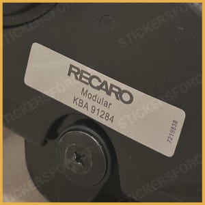 Puede incluir: Una pegatina rectangular plateada con la palabra "RECARO" en negrita y en negro. Debajo, el texto dice "Modular" y "KBA 91284". La pegatina está adherida a una superficie negra, con un tornillo visible debajo.