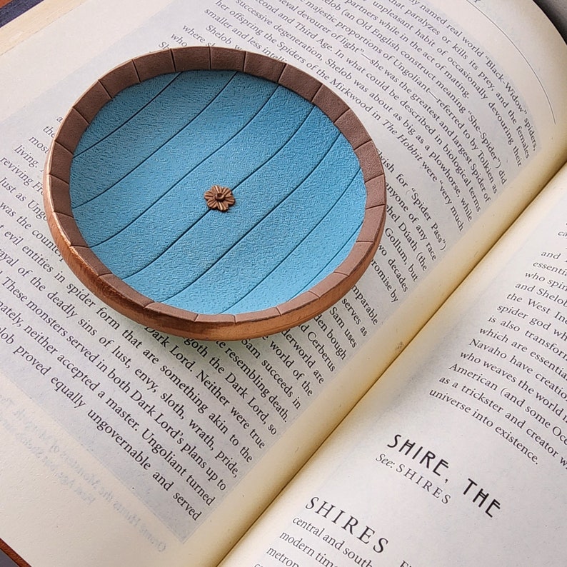 Pu&ograve; includere: Primo piano di un piccolo piatto decorativo rotondo, blu e marrone con un bordo rialzato. Il piatto &egrave; appoggiato su un libro aperto con testo sulle ragnatele.