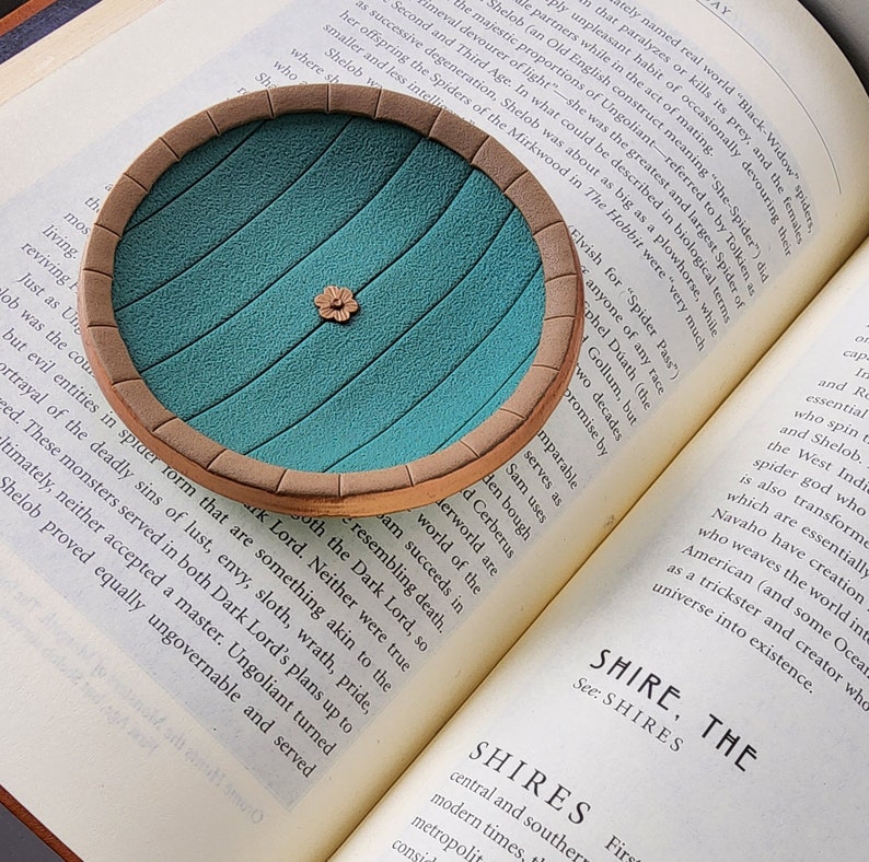 Pu&ograve; includere: Un sottobicchiere in ceramica blu-verde e marrone con un bordo rialzato e un piccolo disegno di un fiore al centro. Il sottobicchiere &egrave; appoggiato su una pagina di un libro sulla Contea dal Signore degli Anelli.