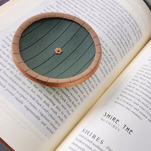 Pu&ograve; includere: Un sottobicchiere circolare verde e marrone con un design rialzato di una porta rotonda con un piccolo fiore al centro. Il sottobicchiere &egrave; appoggiato su un libro con testo sulle ragnatele.
