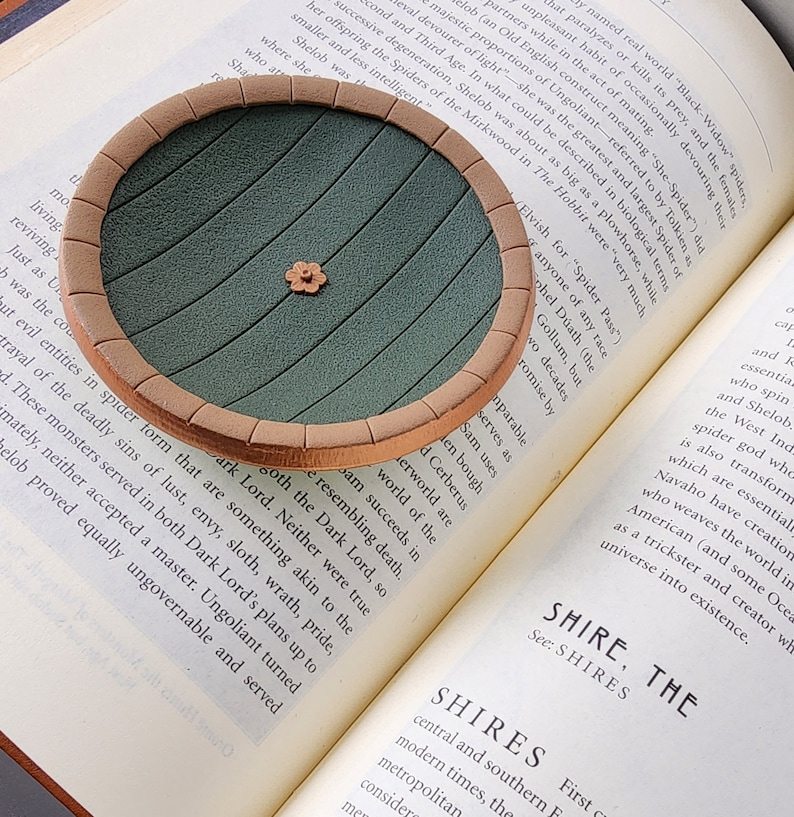 Pu&ograve; includere: Un sottobicchiere circolare verde e marrone con un design rialzato di una porta rotonda con un piccolo fiore al centro. Il sottobicchiere &egrave; appoggiato su un libro con testo sulle ragnatele.