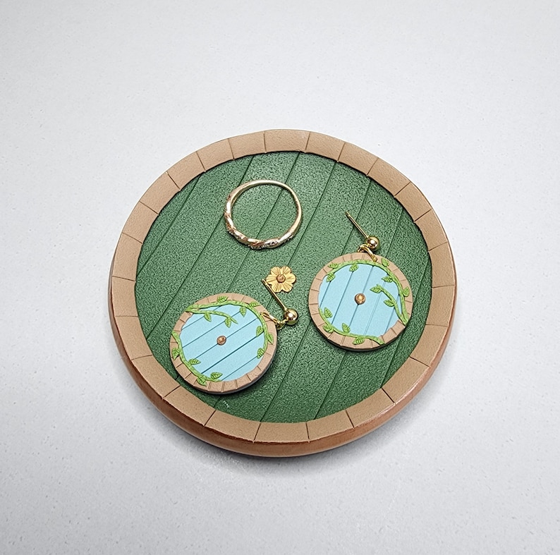 Pu&ograve; includere: Un piatto circolare verde e marrone con un anello d'oro e due orecchini a forma di porta blu. Gli orecchini hanno viti verdi e fiori gialli.