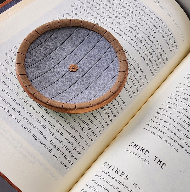 Pu&ograve; includere: Un primo piano di un sottobicchiere rotondo in ceramica grigia con un bordo marrone. Il sottobicchiere ha una superficie testurizzata con linee che lo attraversano. Il sottobicchiere &egrave; appoggiato su un libro con testo sul personaggio di "Shelob" de Il Signore degli Anelli.