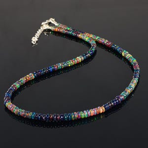 Puede incluir: Un collar de cuentas de ópalo multicolor con un cierre plateado. Las cuentas muestran una gama de colores, incluyendo azules, verdes, naranjas y morados. El collar se muestra sobre una superficie negra.