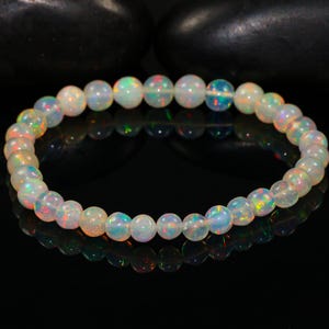 Peut inclure: Un bracelet de perles d'opale iridescentes. Les perles sont principalement blanches avec des éclats de bleu, de vert et d'orange. Le bracelet est présenté sur une surface noire réfléchissante.