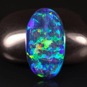 May include: An oval-shaped opal gemstone with a dark blue base color. The opal displays vibrant flashes of green, blue, purple, and orange, creating a colorful, iridescent effect. The gemstone is set against a dark background, highlighting its brilliance.