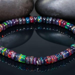 Peut inclure: Un bracelet composé de petites perles d'opale rondes et iridescentes. Les opales présentent une gamme de couleurs, notamment des bleus, des verts, des rouges et des violets. Le bracelet est présenté sur une surface noire réfléchissante.