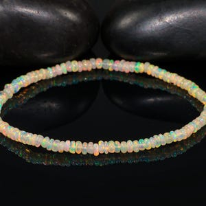 Peut inclure: Un bracelet délicat composé de petites perles d'opale iridescentes. Les perles affichent une gamme de couleurs, dont le blanc, le jaune, le vert et le rose. Le bracelet est présenté sur une surface noire réfléchissante, avec deux pierres sombres et arrondies en arrière-plan.